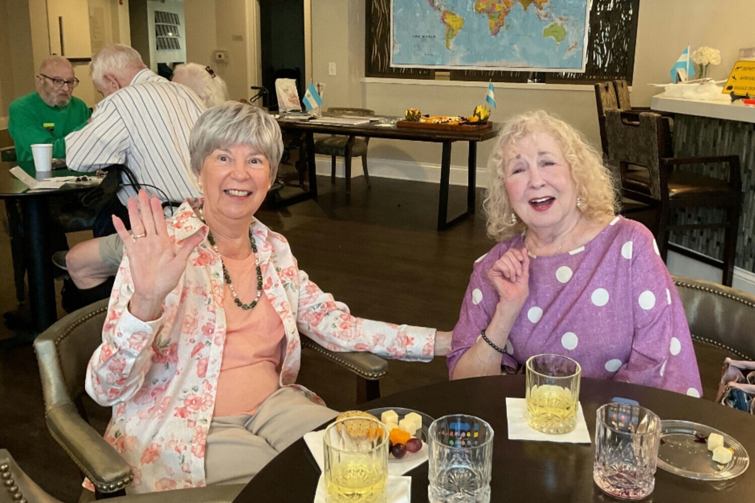 Two residents laugh and wave cheerfully while enjoying a meal together in the dining area at Arbor Terrace Hamilton Mill, a senior living community in Dacula, GA, in a warm and social atmosphere.