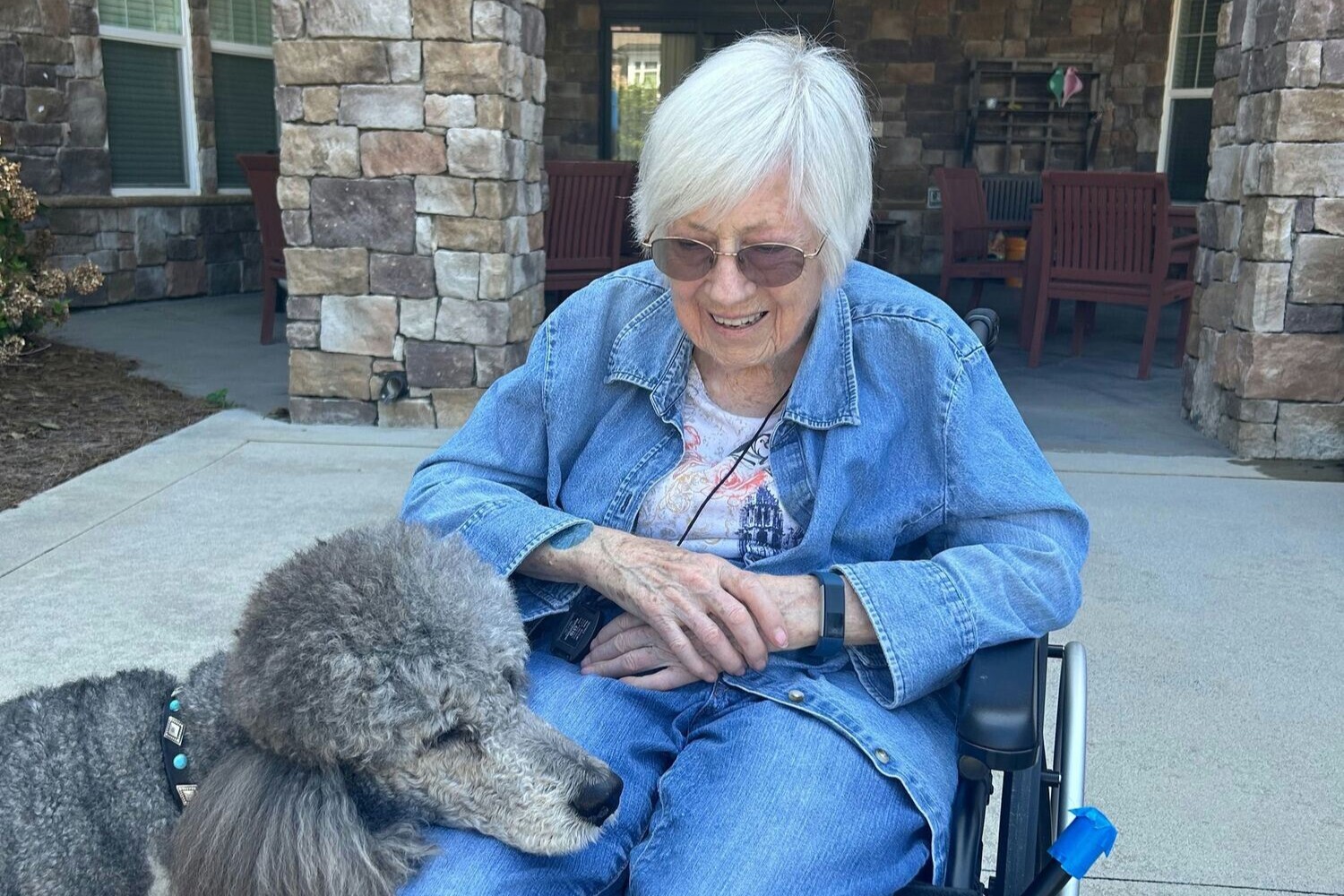 A resident enjoys a sweet moment outdoors with a fluffy grey dog at Arbor Terrace Hamilton Mill, a pet-friendly senior living community in Dacula, GA, smiling warmly on a sunny patio.