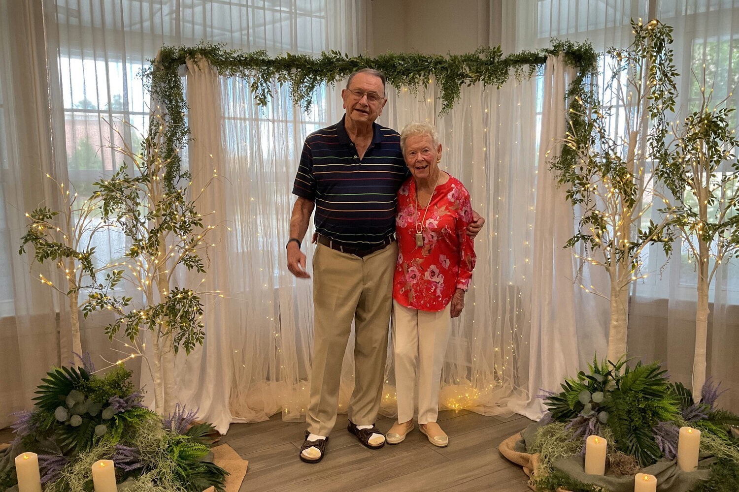 A senior couple standing together and smiling in front of a beautifully decorated floral and greenery backdrop — capturing the warmth and connection found in retirement communities Dacula GA.