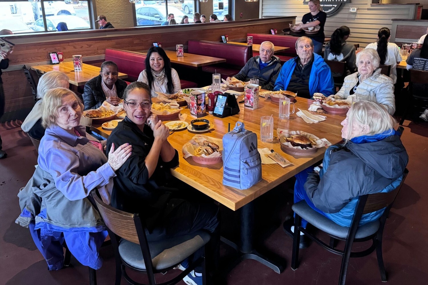 A lively group of seniors and staff sharing a meal together at a restaurant, laughing and enjoying each other's company — the kind of social connection at the heart of senior living Dacula Georgia.