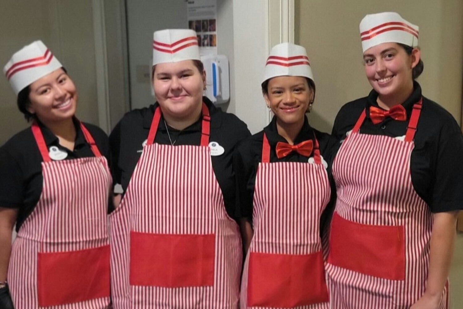 Four smiling dining team members in matching red-striped aprons and white hats posing together — the dedicated staff behind the senior living with gourmet dining Dacula GA residents enjoy every day.