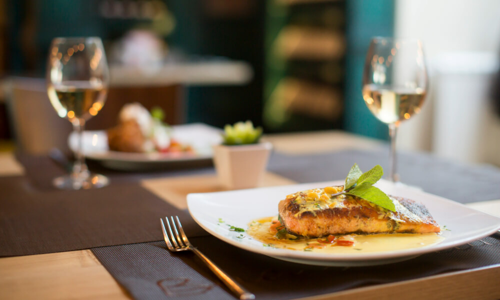 A beautifully plated salmon dish with fresh herbs and sauce, served alongside wine glasses on an elegantly set table — a taste of the senior living with gourmet dining Dacula GA residents savor daily.