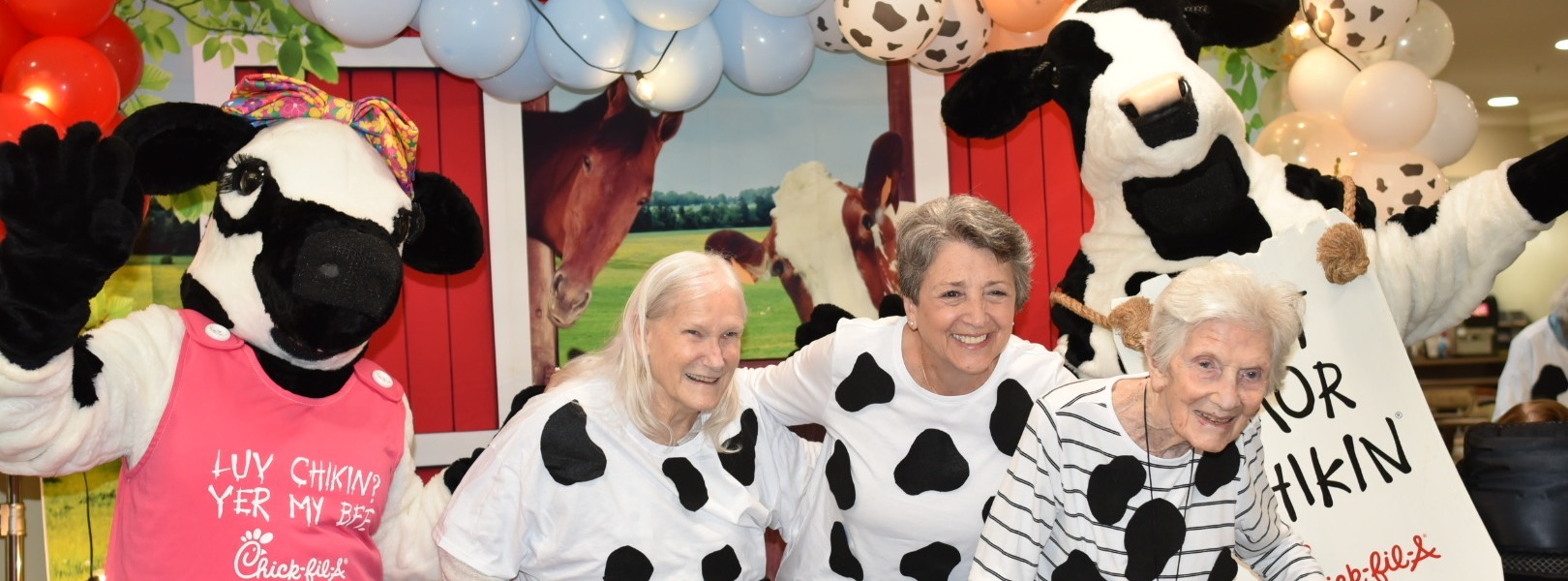 Three smiling seniors pose with cow mascots amid festive balloons and farm-themed decor during a lively celebration at this memory care community near Hamilton Mill.