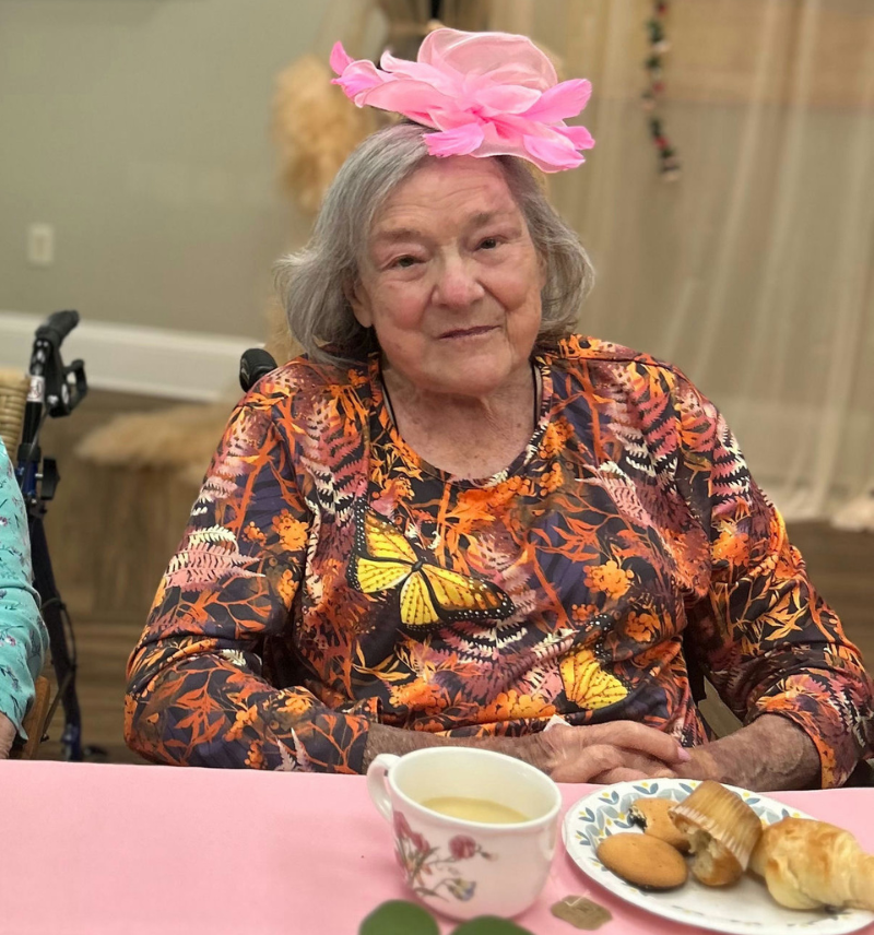 A senior resident smiles warmly while enjoying tea and snacks at a table, wearing a cheerful pink flower hat at this Bridges program Dacula GA community.