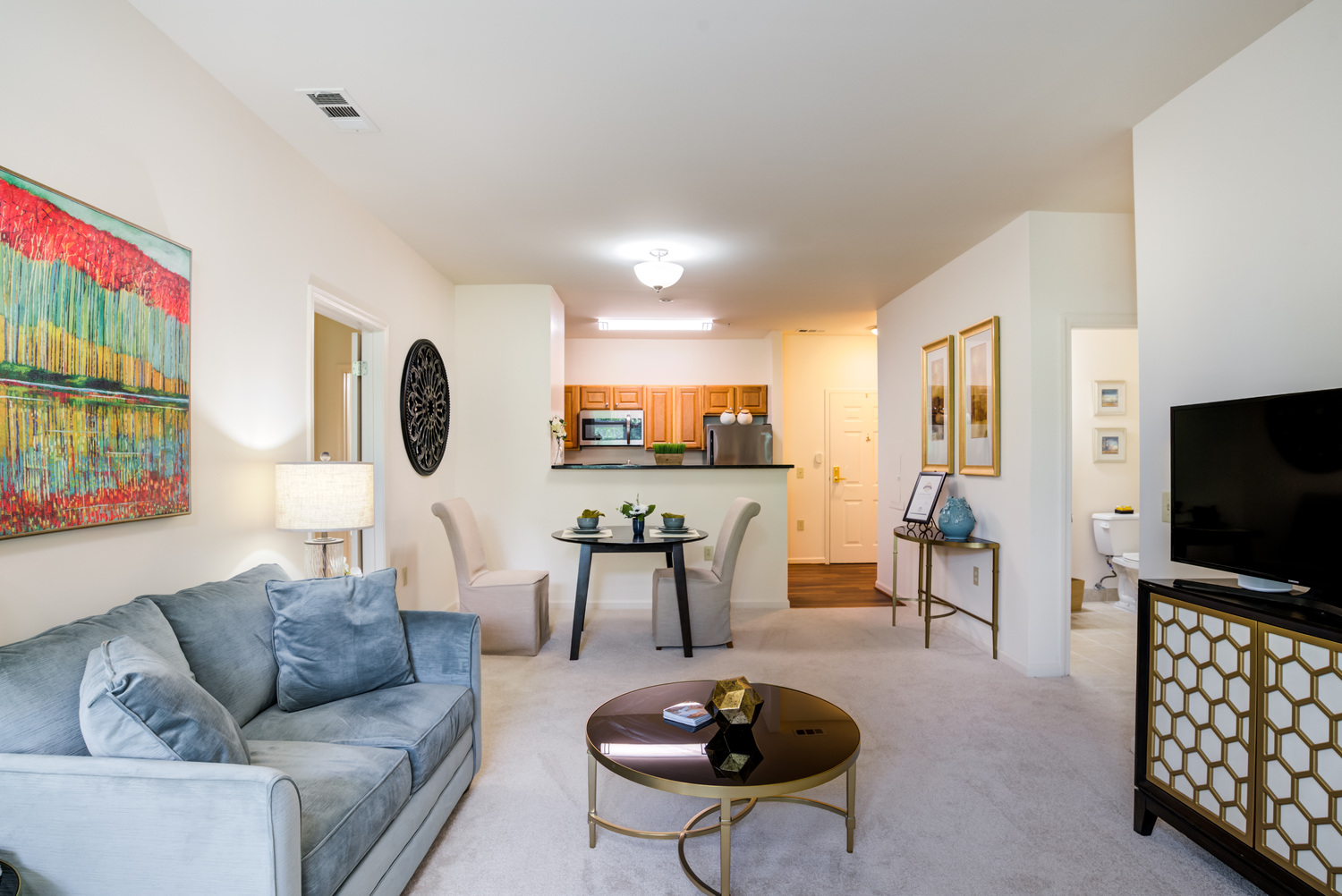 An open-concept apartment living area at Arbor Terrace Herndon with a sofa, wall art, and a view into the kitchen, offering a welcoming home for seniors in Herndon, VA.