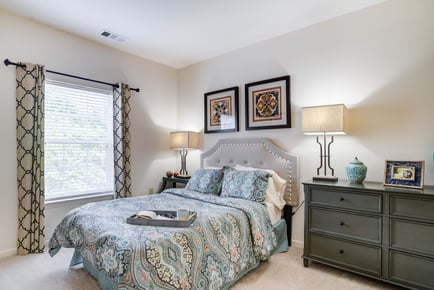 A welcoming independent living bedroom in Herndon VA, featuring a tufted upholstered headboard, a blue paisley comforter, patterned curtains, a dark wood dresser, warm bedside lamps, and two framed art pieces above the bed.