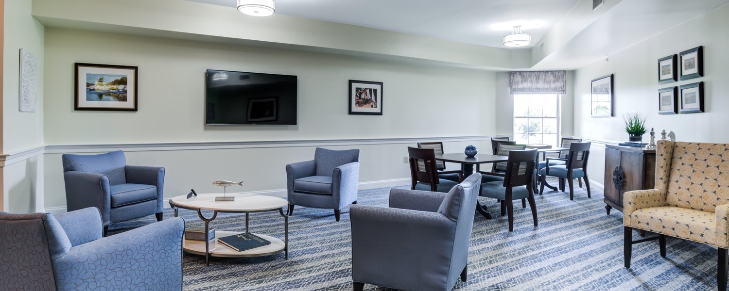 A comfortable common lounge features plush blue armchairs, a wall-mounted TV, and tasteful artwork, showcasing the inviting independent living Herndon VA spaces at Arbor Terrace Herndon, a warm senior living Herndon Virginia community.
