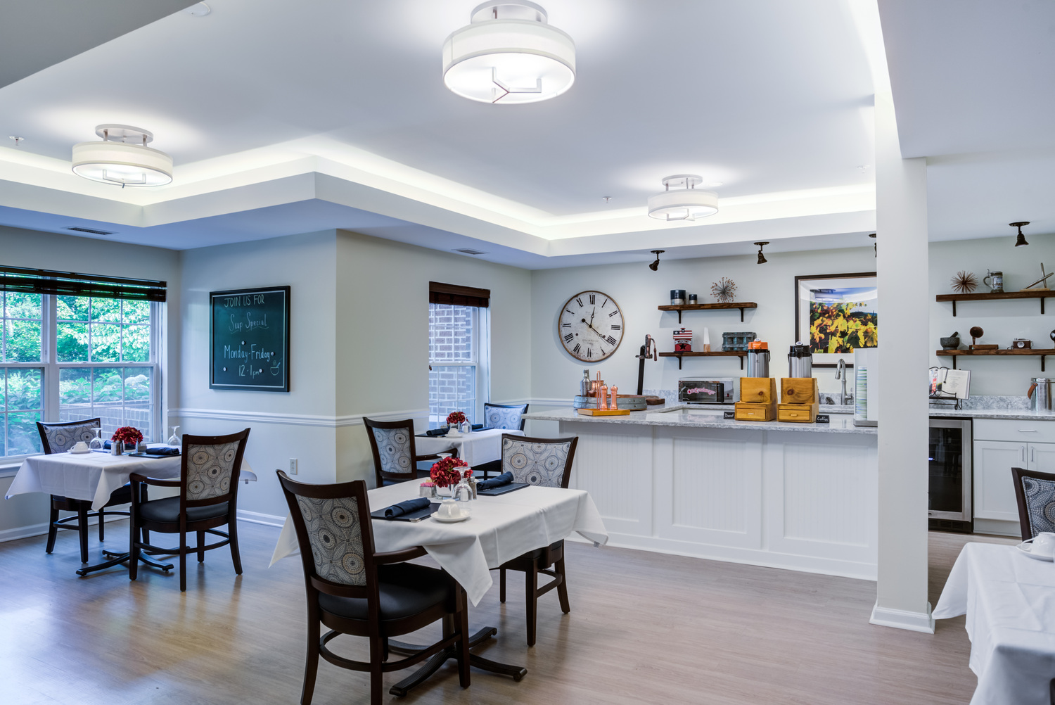A bright dining area at Arbor Terrace Herndon featuring white-topped tables, dark wood chairs, a wall clock, and open shelving, supporting a vibrant senior living experience in Herndon, VA.