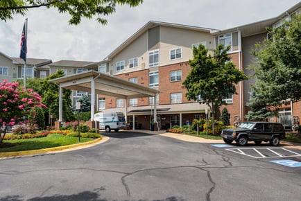The welcoming exterior of Arbor Terrace Herndon, a well-landscaped senior living community in Herndon, VA, featuring a brick facade, lush greenery, and a covered entrance.