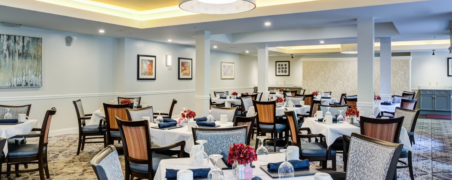 A spacious, elegantly set dining room at Arbor Terrace Herndon with round tables, upholstered chairs, and warm lighting, offering restaurant-style dining for seniors in Herndon, VA.