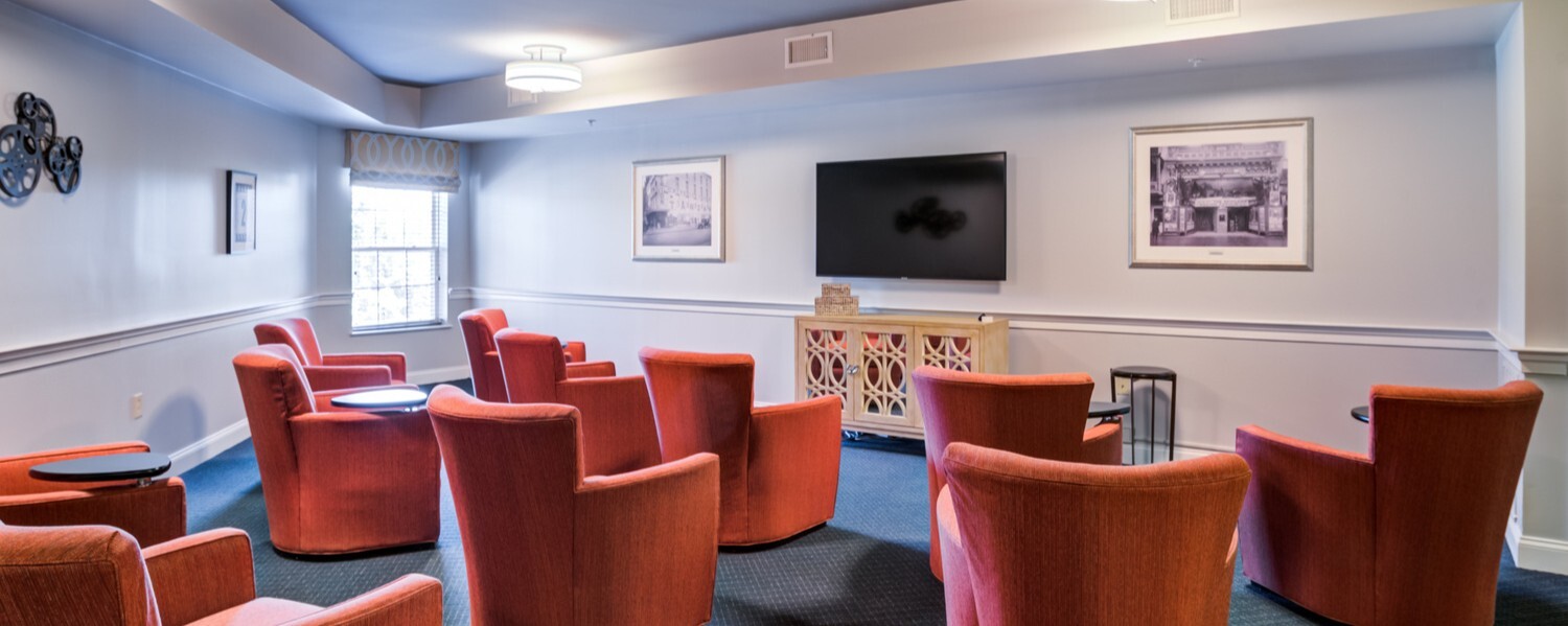 A cozy private theater at Arbor Terrace Herndon with rows of plush red chairs and a wall-mounted screen, providing entertainment and enrichment for residents of this Herndon, VA retirement community.