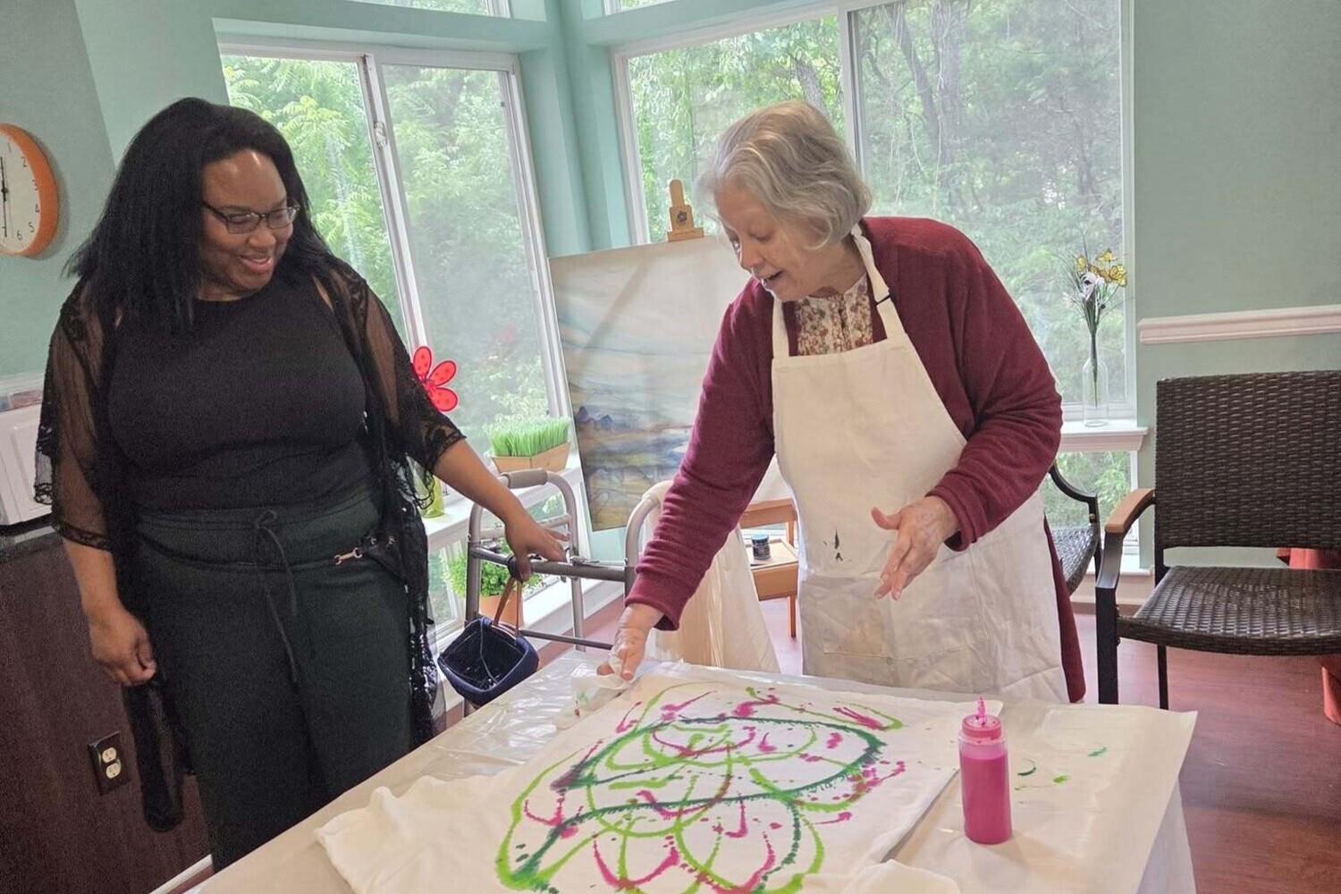 Two women share a creative moment during a colorful paint pouring activity in a sunny art studio, reflecting the vibrant senior living lifestyle and creative spirit found throughout this senior living community.