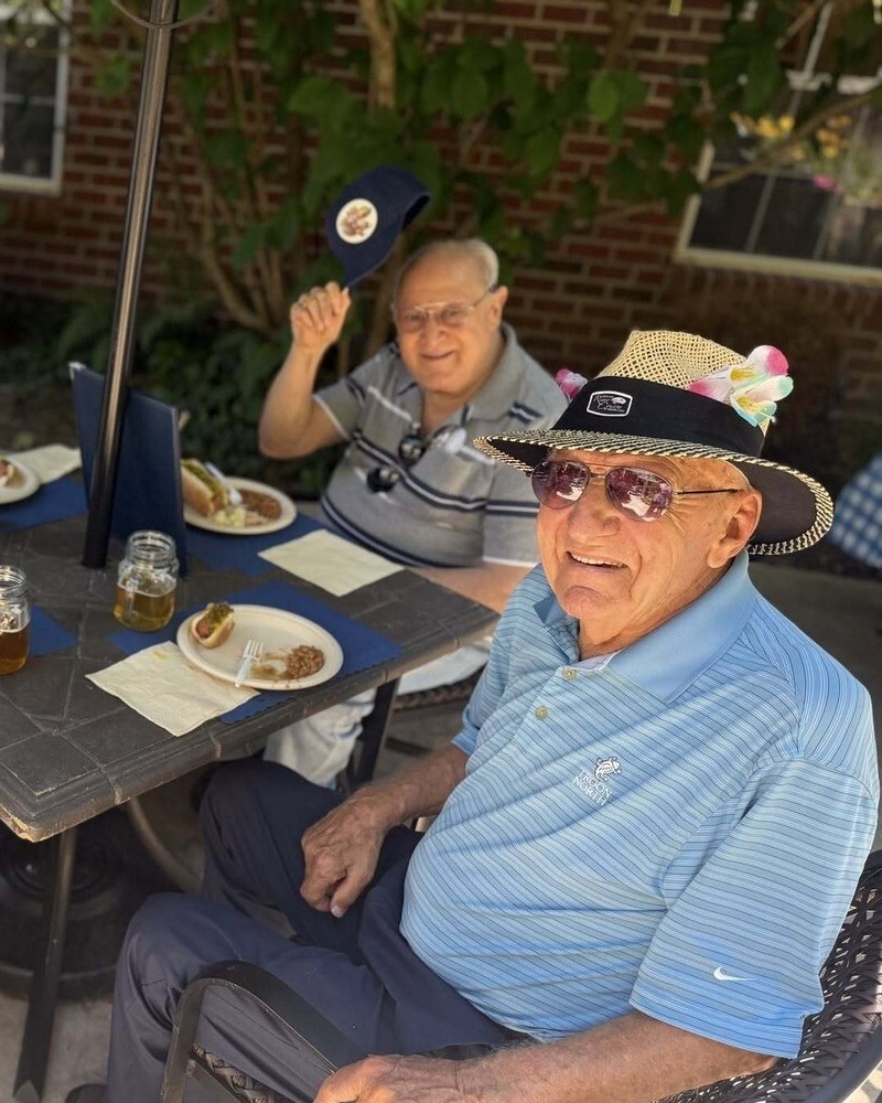 Two residents share a sunny outdoor cookout, smiling in playful summer hats over plates of hot dogs, capturing the joyful senior living with dining and activities Herndon at Arbor Terrace Herndon, a lively senior living Herndon Virginia community.