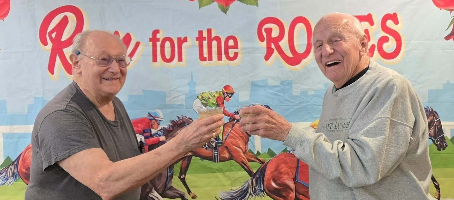 Two residents share a cheerful toast in front of a festive Run for the Roses banner featuring horse racing artwork, celebrating together at Arbor Terrace Herndon, a senior living Herndon Virginia community that embraces an active senior living Herndon VA spirit.