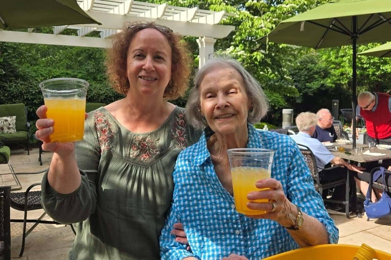 Two women raise their glasses in a cheerful toast on a sunny, green patio, capturing a warm social moment and the vibrant senior living lifestyle enjoyed throughout this senior living community.