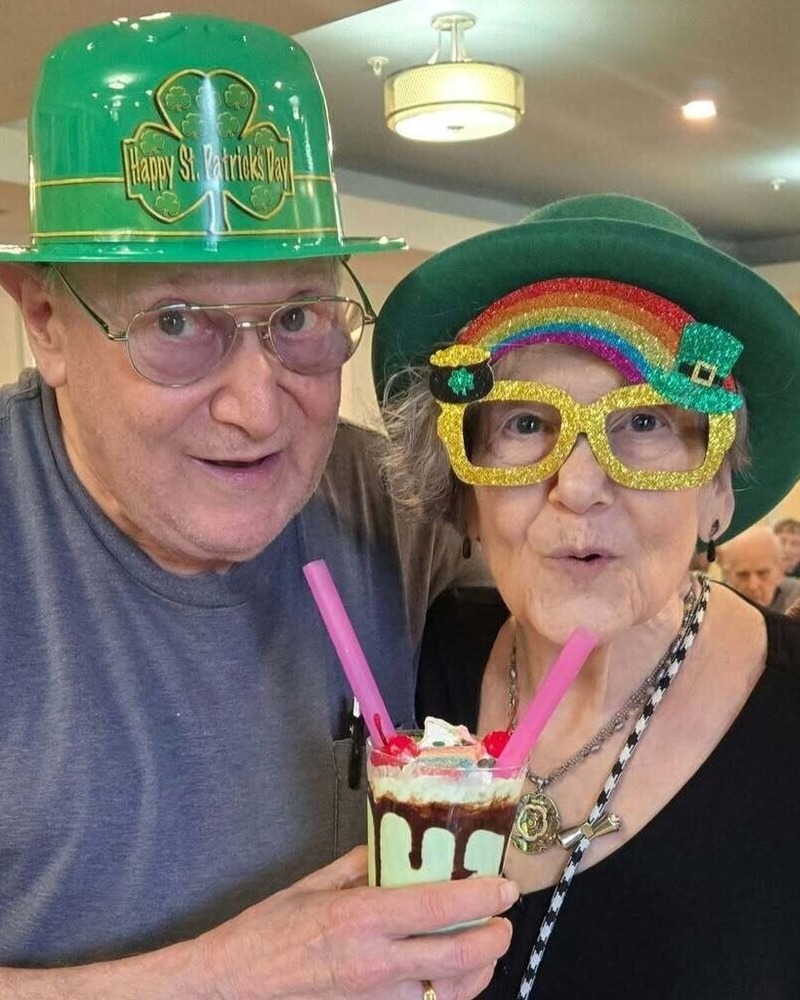 Two residents celebrate St. Patrick's Day in festive green hats and colorful accessories, sharing a whimsical whipped-cream treat, capturing the joyful senior living with dining and activities Herndon spirit at Arbor Terrace Herndon, a vibrant senior living Herndon Virginia community.