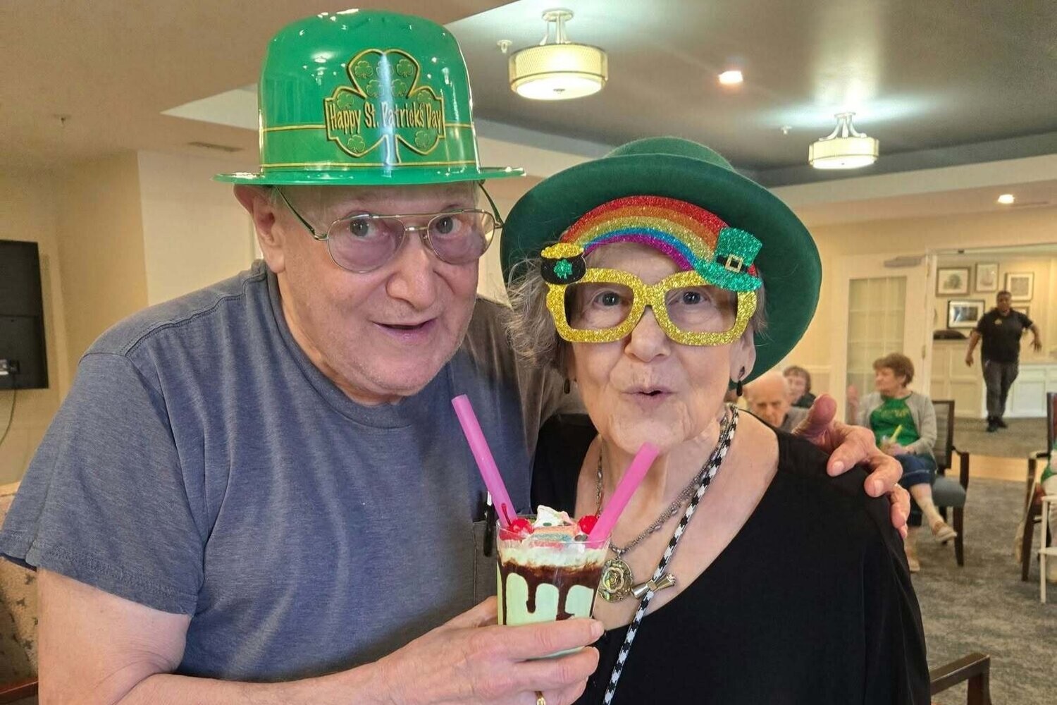 Two residents celebrate St. Patrick's Day in festive green hats and colorful accessories, sharing a whimsical whipped-cream treat, capturing the joyful senior living with dining and activities Herndon spirit at Arbor Terrace Herndon, a vibrant senior living Herndon Virginia community.