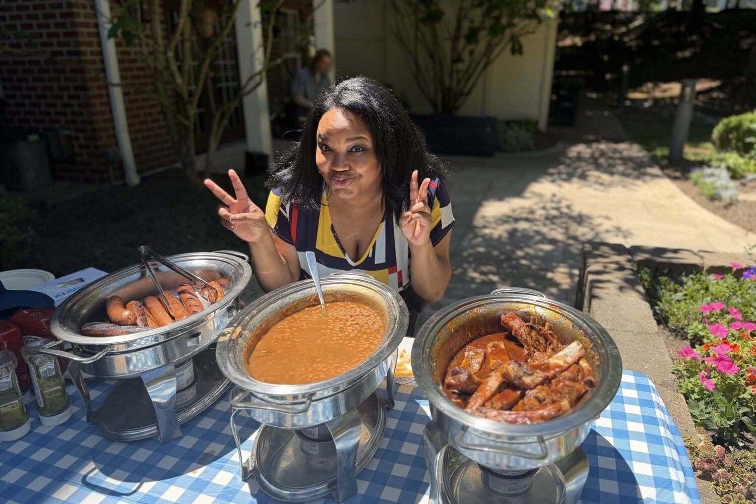 A cheerful Arbor Company team member gives a playful peace sign while serving a festive outdoor meal from chafing dishes, capturing the lively spirit of senior living community life.
