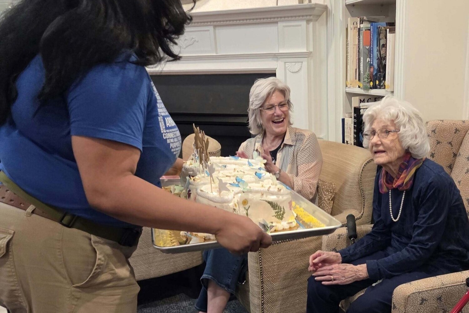 A staff member presents a beautifully decorated cake to two smiling residents by a cozy fireplace, celebrating a special occasion at Arbor Terrace Herndon, a senior living Herndon Virginia community known for its senior living with dining and activities Herndon.