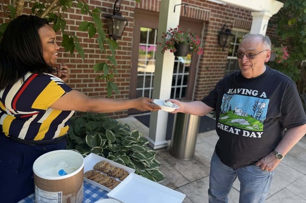 A smiling staff member shares a treat with a resident on a sunny patio at Arbor Terrace Herndon, capturing the active senior living Herndon VA lifestyle within this welcoming senior living Herndon Virginia community.