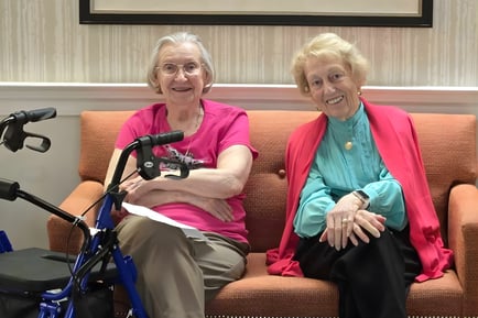 Two women sit together smiling on an orange sofa in a cozy common area at Arbor Terrace Herndon, illustrating the warm and welcoming senior living community in Herndon, Virginia.