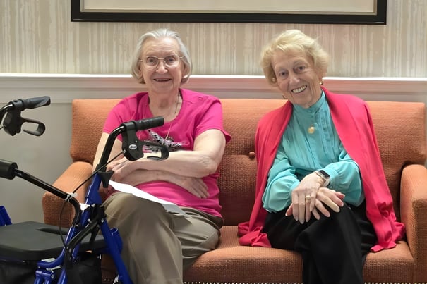 Two women sit together smiling on an orange sofa in a cozy common area at Arbor Terrace Herndon, illustrating the warm and welcoming senior living community in Herndon, Virginia.