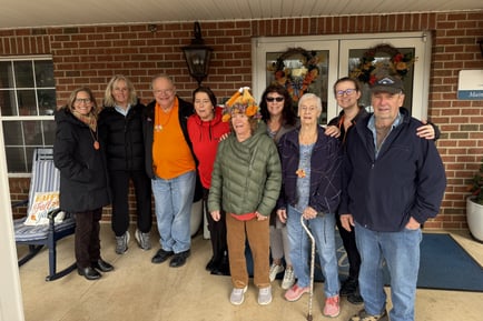 A group of approximately ten adults of varying ages posing together outdoors on a porch or entryway in front of a brick building, smiling for a group photo. 