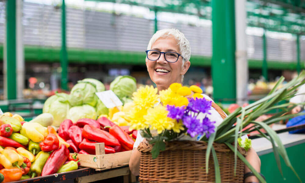 A smiling woman browses colorful fresh produce and flowers at an outdoor farmers market, reflecting the vibrant, walkable lifestyle available near senior living in Herndon, VA.