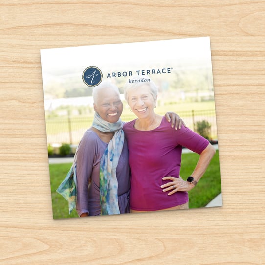 Square photo of two smiling senior women embracing outdoors, with Arbor Terrace Herndon logo above, displayed on a printed card against a wood-grain background.