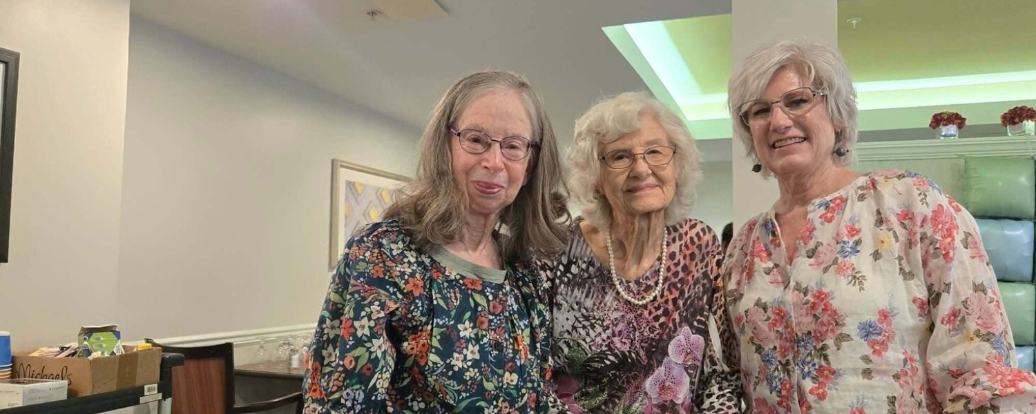 Three smiling women share a warm, joyful moment together in a bright room, reflecting the welcoming friendships and vibrant senior living lifestyle found throughout this senior living community.