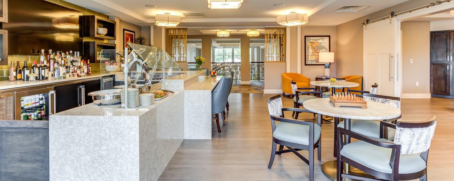Round tables with patterned chairs and a game board sit beside a bar with shelves of bottles and a stone counter with stacked plates in a bright space at Arbor Terrace Highland Park, one of the top senior living communities Lake County IL for an active senior lifestyle Highland Park IL.