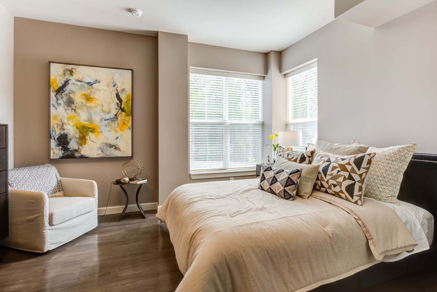 A stylish, comfortable bedroom at this assisted living community in Highland Park IL, featuring warm wood flooring, a plush upholstered bed with layered neutral pillows, a bold abstract painting, a cozy armchair, and a bright window with soft natural light.