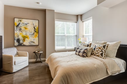 A stylish, comfortable bedroom at this assisted living community in Highland Park IL, featuring warm wood flooring, a plush upholstered bed with layered neutral pillows, a bold abstract painting, a cozy armchair, and a bright window with soft natural light.