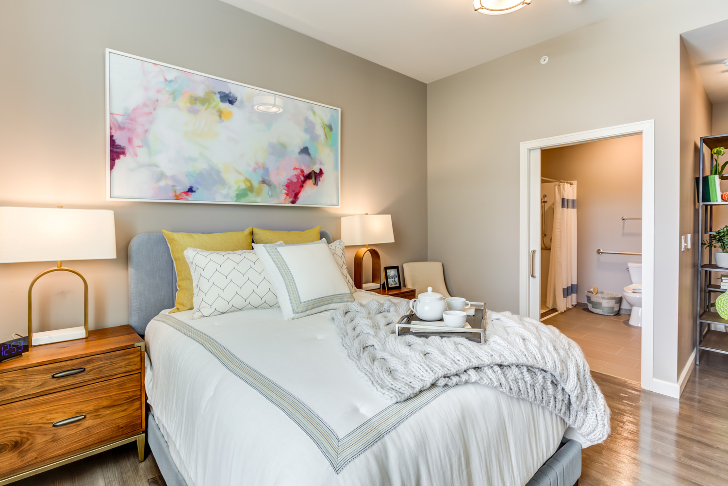 Bright bedroom with a gray upholstered headboard, layered white and yellow pillows, wood nightstands with brass lamps, and an ensuite bathroom, part of the senior apartments near downtown Highland Park at our senior living community.