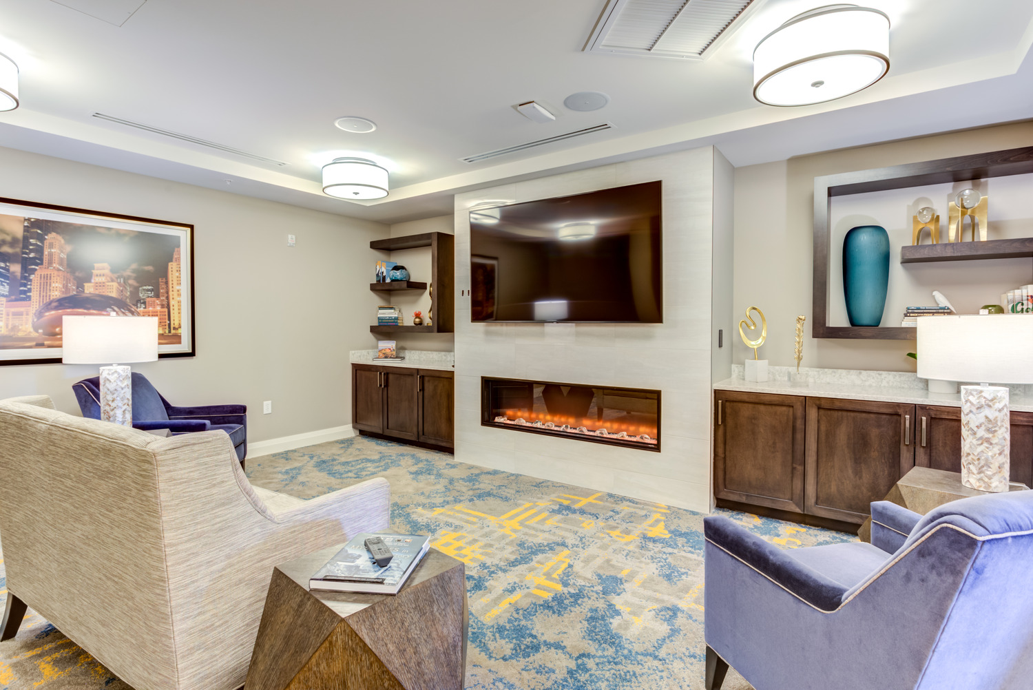 Cozy lounge with a linear fireplace, mounted TV, plush armchairs, and a city skyline print, part of the senior living community in Highland Park, IL, where residents can relax and unwind.