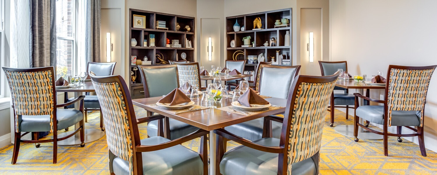 Wood tables set with napkins and glassware are surrounded by patterned upholstered chairs in the bright dining room at Arbor Terrace Highland Park, with large windows and built-in shelves, a welcoming place to gather for senior living Highland Park IL and senior care Highland Park Illinois.