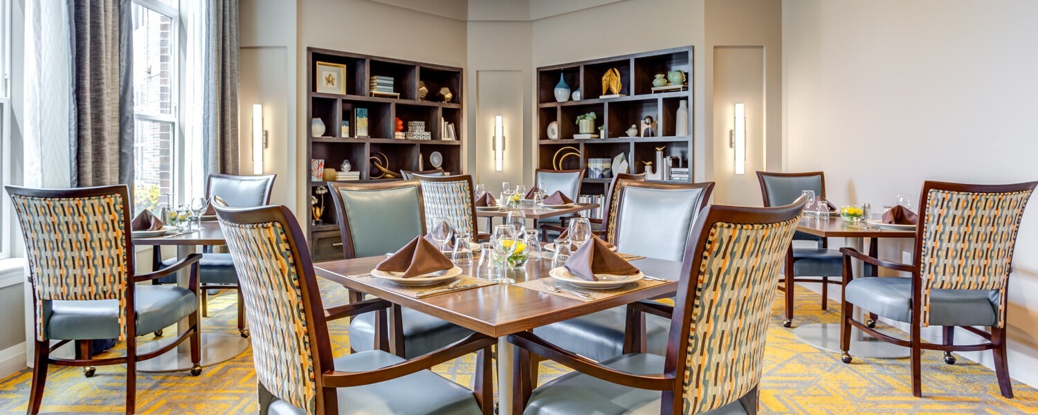 An elegant dining room with wooden tables set with folded napkins and glassware, upholstered chairs with woven backs, built-in shelves, and tall draped windows, a welcoming space at a Highland Park IL assisted living community among top senior living communities Lake County IL.