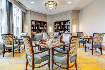 A dining room at Arbor Terrace Highland Park features tables set with folded napkins, wine glasses, and floral centerpieces, surrounded by patterned chairs, built-in display shelving, and a bold yellow-and-gray carpet. It reflects the resort-style senior living in Highland Park, IL.