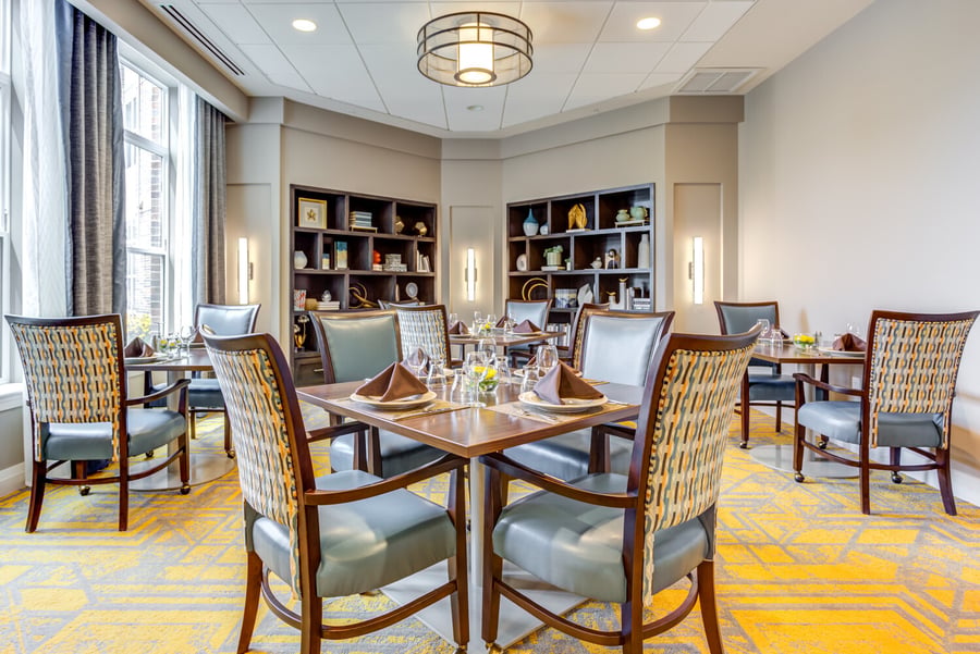 A dining room at Arbor Terrace Highland Park features tables set with folded napkins, wine glasses, and floral centerpieces, surrounded by patterned chairs, built-in display shelving, and a bold yellow-and-gray carpet. It reflects the resort-style senior living in Highland Park, IL.