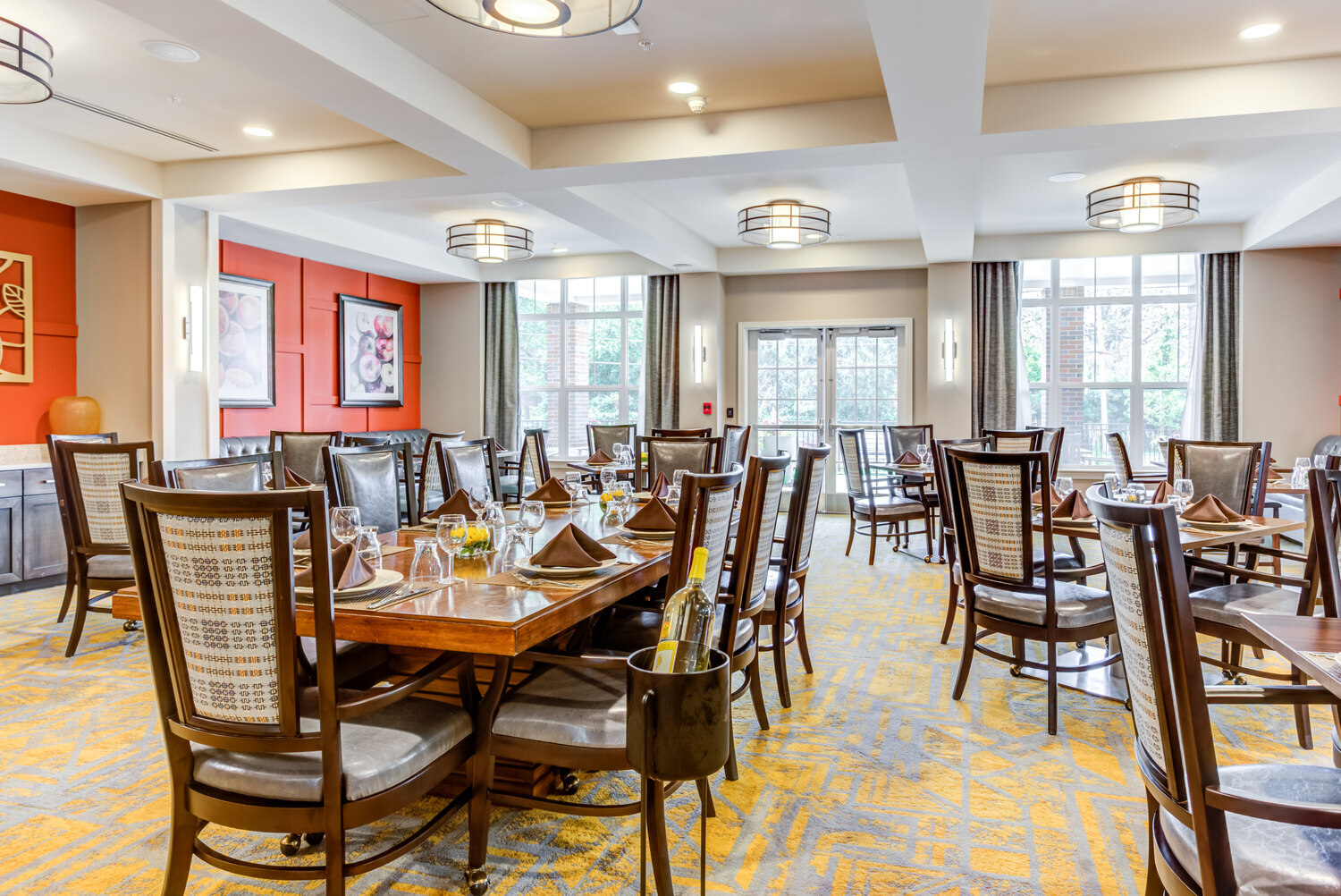 Sunlit dining room with wood tables set for a meal, patterned carpet, and large windows, part of the senior living community in Highland Park, IL, offering a bright space to gather.