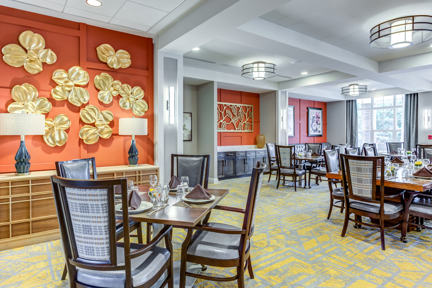 Vibrant dining room with an orange accent wall, gold leaf art, and set tables, part of the senior living community in Highland Park, IL, offering a welcoming space to dine.