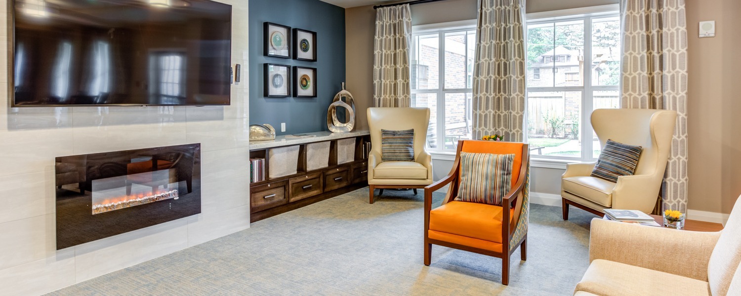 A cozy lounge at Arbor Terrace Highland Park features a linear fireplace beneath a wall-mounted TV, cream wingback chairs, an orange armchair, and tall windows with patterned curtains, a comfortable spot to relax for high-end assisted living North Shore and senior living Highland Park IL.