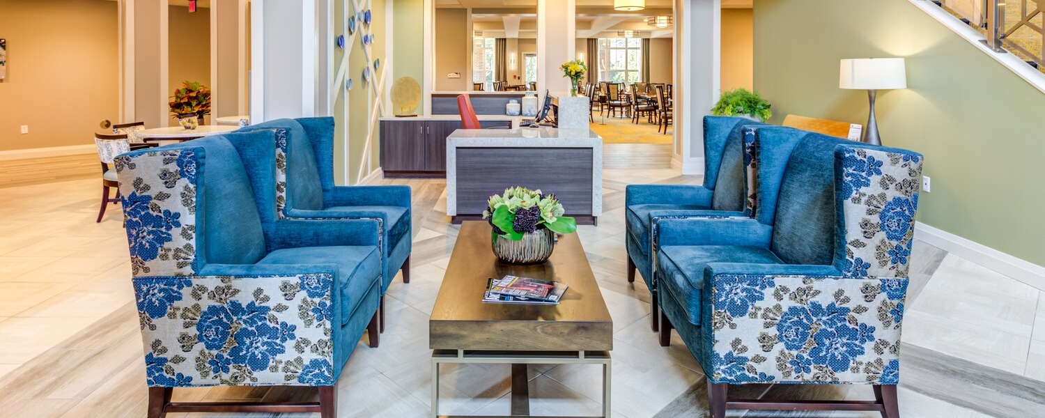 The welcoming lobby at Arbor Terrace Highland Park features blue floral wingback chairs around a wood coffee table with fresh flowers, opening to a bright dining area beyond. A comfortable gathering spot for senior living Highland Park IL and boutique assisted living Highland Park.