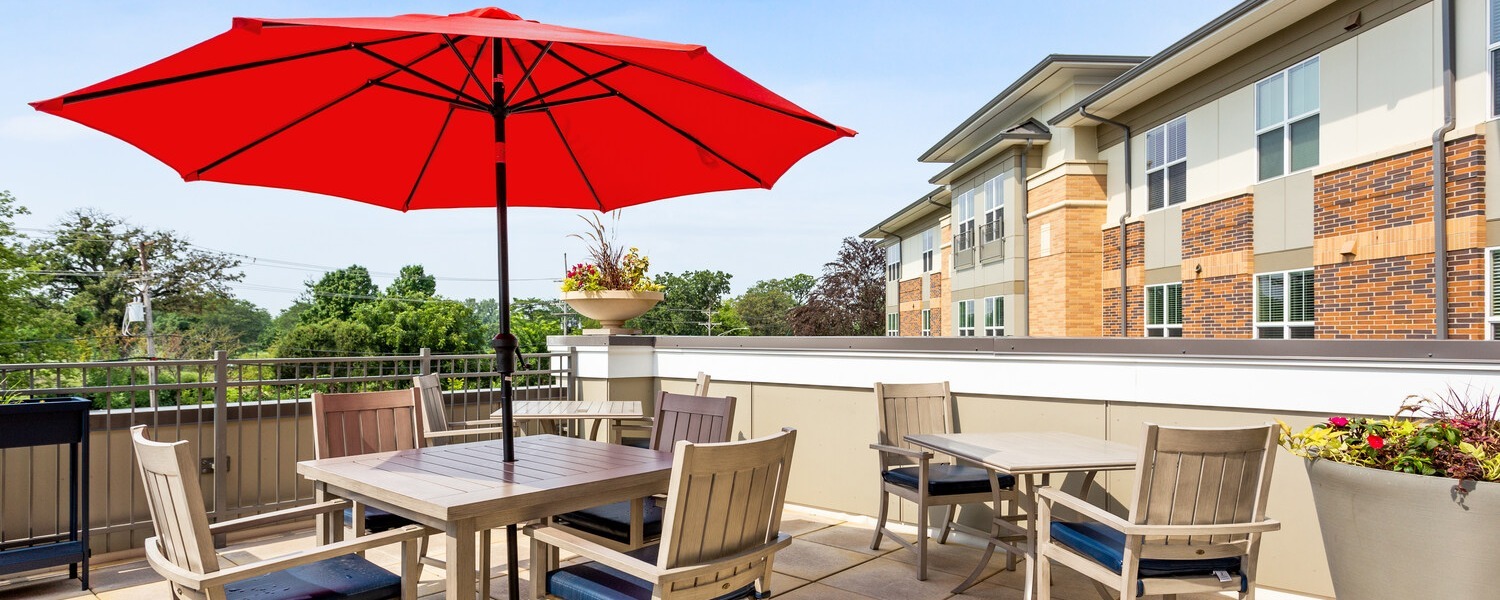 Sunny outdoor patio with a red umbrella, wooden chairs, and blooming flowers, part of the active senior lifestyle Highland Park IL residents enjoy at this senior living Highland Park IL community.