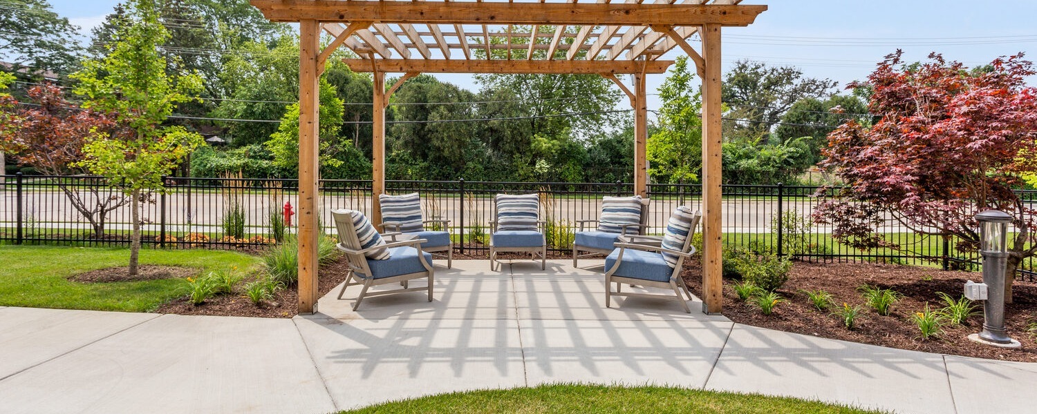 A wooden pergola shades a sunny patio with cushioned armchairs, surrounded by landscaped garden beds, a red-leaf maple, and leafy trees along a paved walkway, an inviting outdoor retreat for nature-friendly senior living Highland Park IL at a Highland Park IL assisted living community.