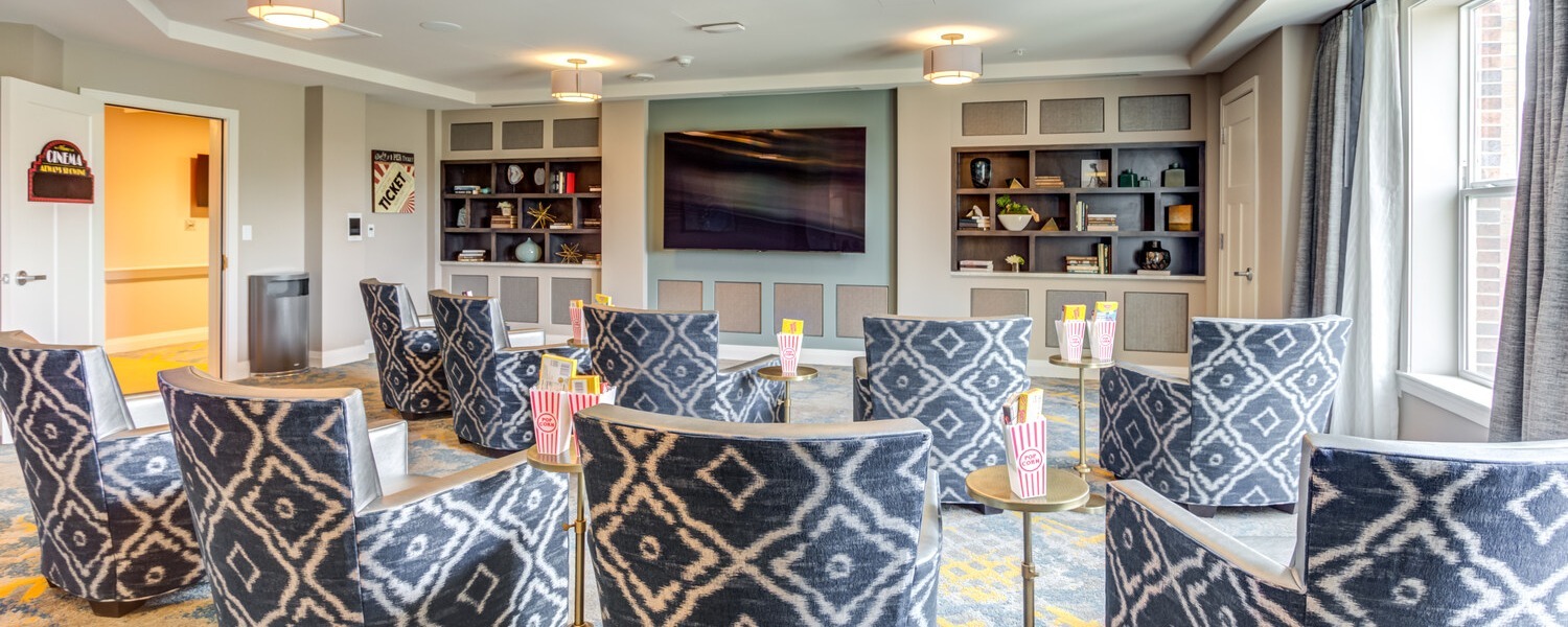 A cozy lounge at Arbor Terrace Highland Park offers navy and white patterned armchairs facing a large wall-mounted TV, with popcorn boxes ready on small side tables, a relaxed spot to gather that reflects an active senior lifestyle Highland Park IL and senior care Highland Park Illinois.