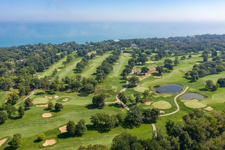 Aerial view of rolling green fairways and sand bunkers bordered by trees, with blue water stretching along the horizon, a scenic backdrop for assisted living Glencoe IL and retirement living near North Shore golf courses.