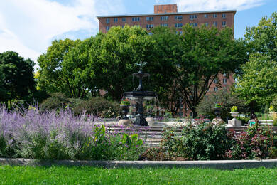 A tiered stone fountain rises among blooming purple flowers and leafy trees in a sunny garden, with a brick building in the distance, a relaxing outdoor spot for nature-friendly senior living Highland Park IL and senior care Highland Park Illinois.