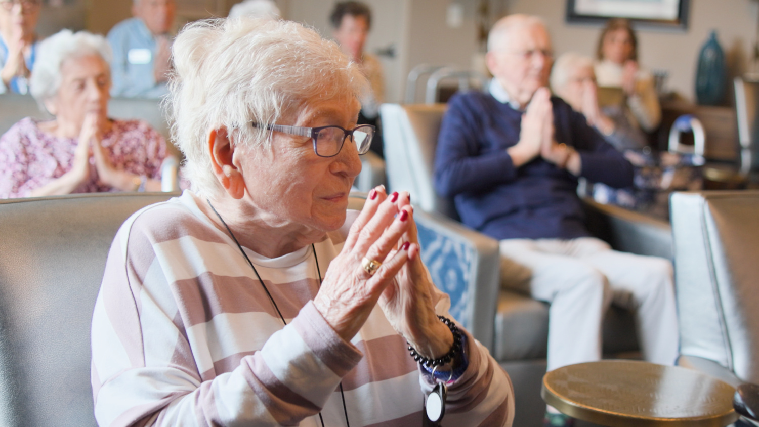 A resident sits with hands clasped in quiet prayer, surrounded by fellow residents doing the same in a bright common area, reflecting a peaceful moment at our senior living Highland Park IL community and its active senior lifestyle Highland Park IL.