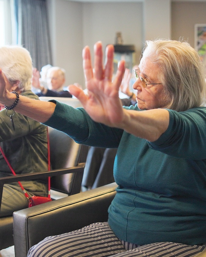A community member in a teal sweater and glasses stretches her arms forward with palms raised as others join in from their seats during a group movement session at Arbor Terrace Highland Park, reflecting wellness-focused senior living North Shore and senior care Highland Park Illinois.